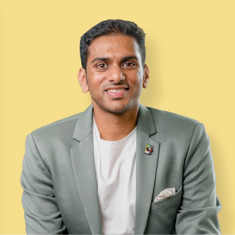 Professional portrait of a smiling man in a sage green blazer and white shirt against a yellow background for business headshot photography.