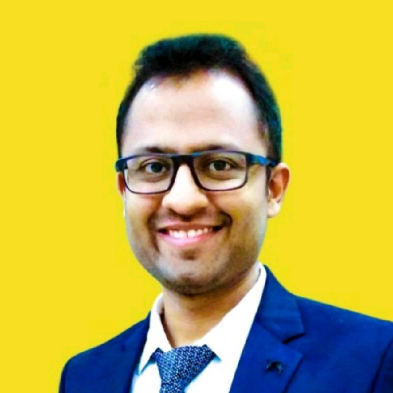 Professional headshot of a smiling man wearing glasses and a blue blazer against a yellow background for corporate or business profile use.