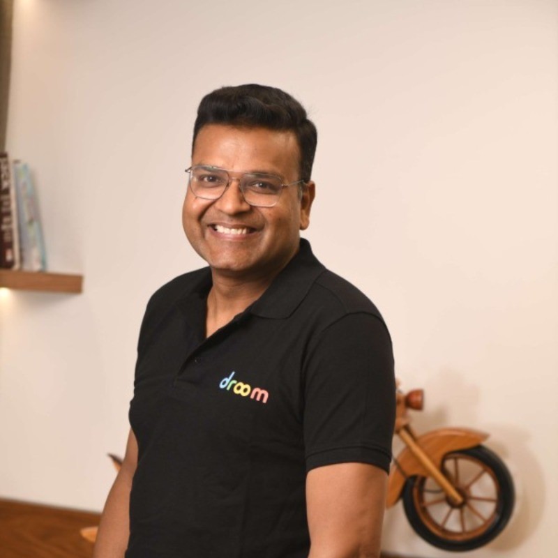 Professional man wearing glasses and a black Droom branded polo shirt smiles at the camera in an indoor office setting with wooden decor.