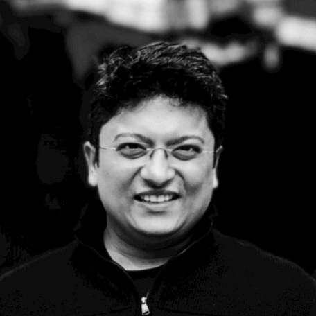 A smiling man with dark hair wearing glasses and a black jacket in a professional black and white portrait photograph.