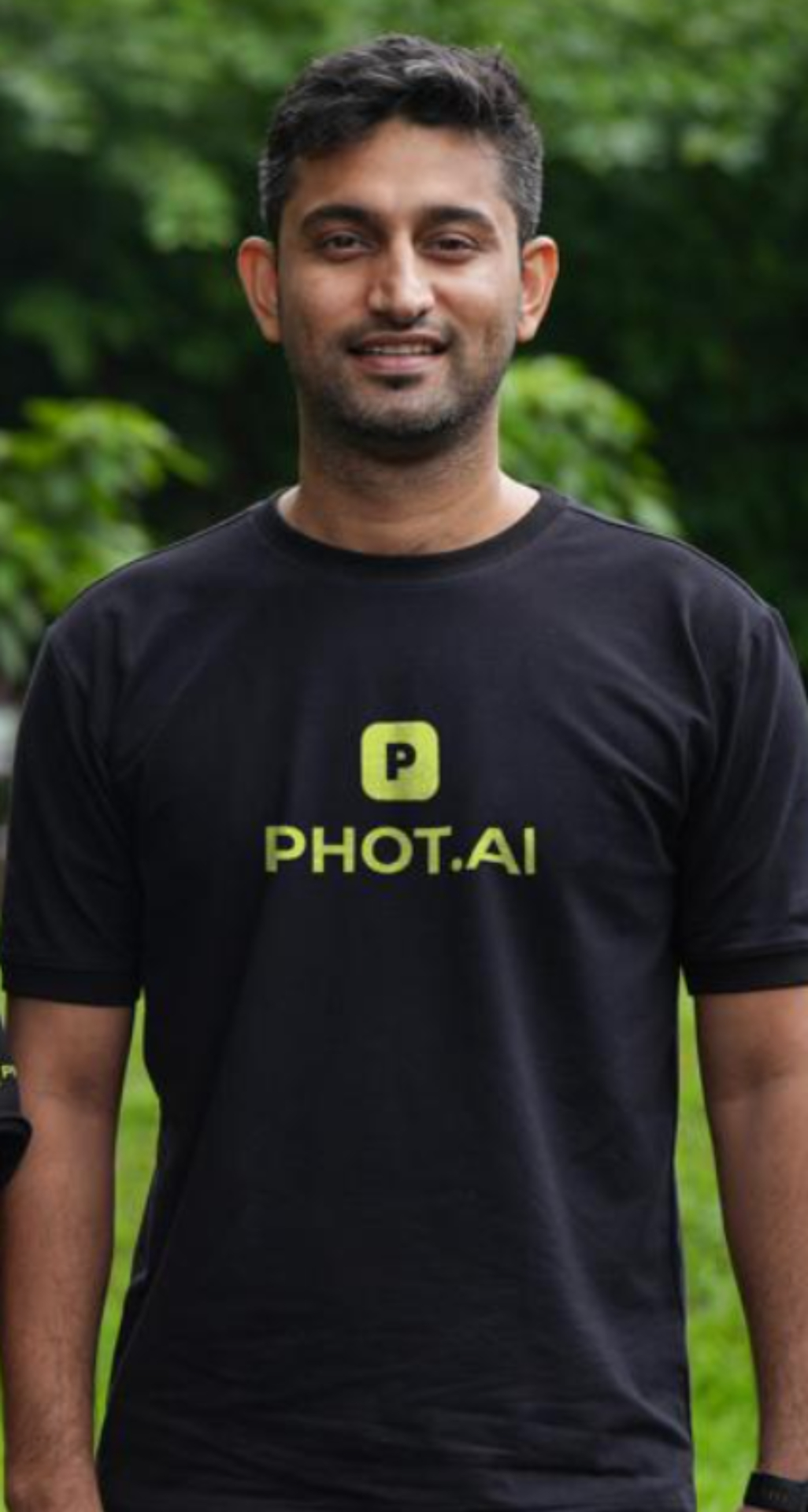 A smiling man with long curly dark hair and glasses wearing a black PHOT.AI t-shirt stands outdoors in front of green foliage.