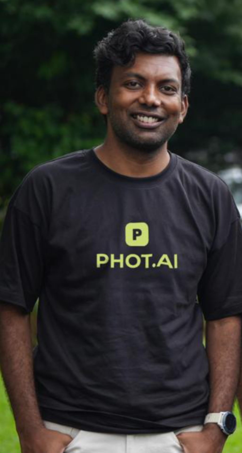 A smiling man wearing a black PHOT.AI branded t-shirt stands outdoors against a blurred green foliage background in a professional headshot portrait.