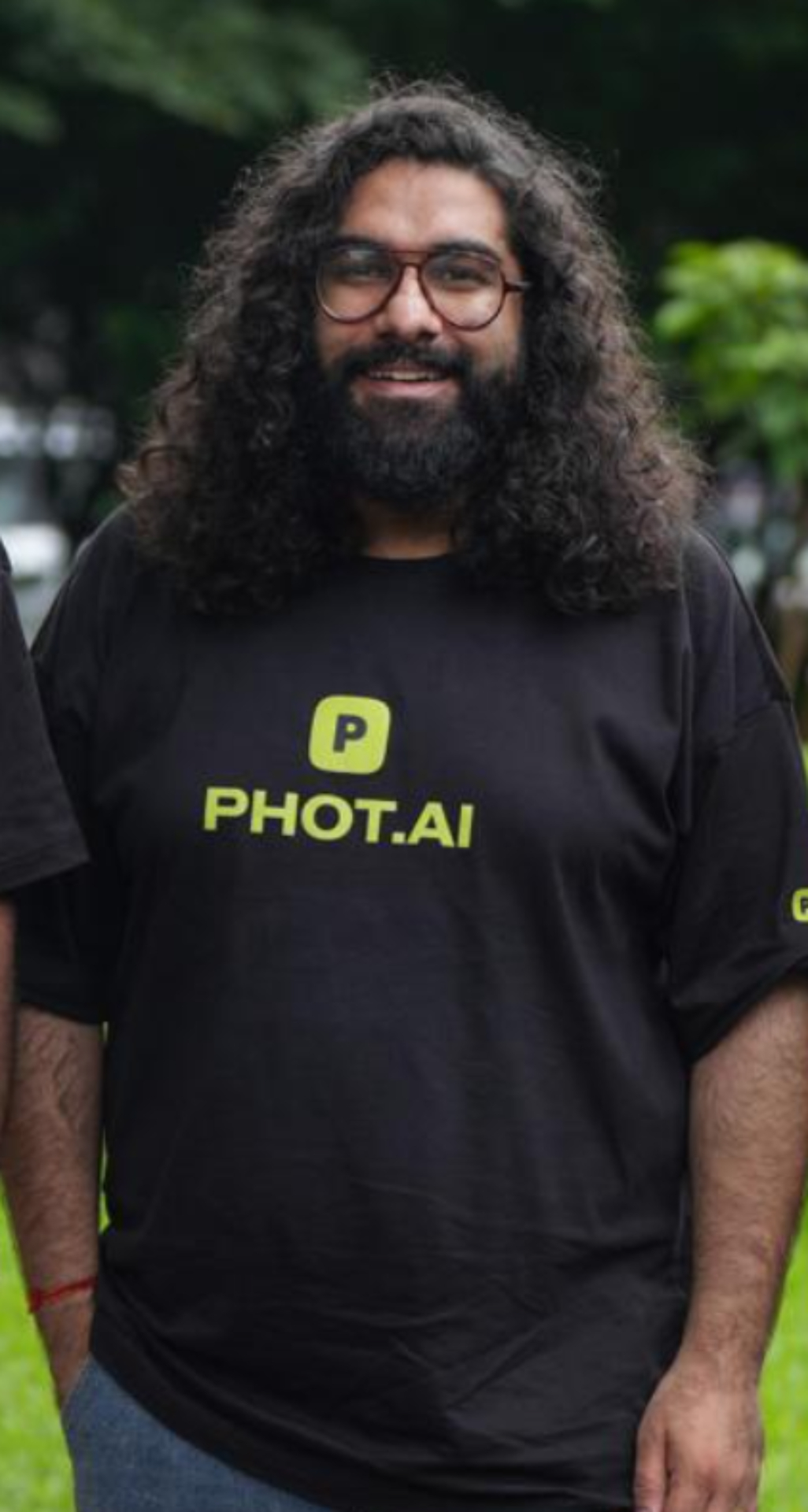 Smiling man wearing black PHOT.AI branded t-shirt standing outdoors with green blurred background in professional portrait photo.
