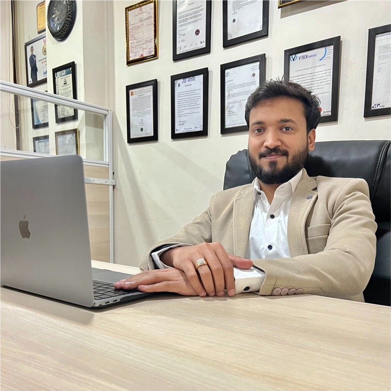 Professional businessman in beige blazer sitting at desk with laptop in modern office workspace decorated with framed certificates and qualifications on wall.