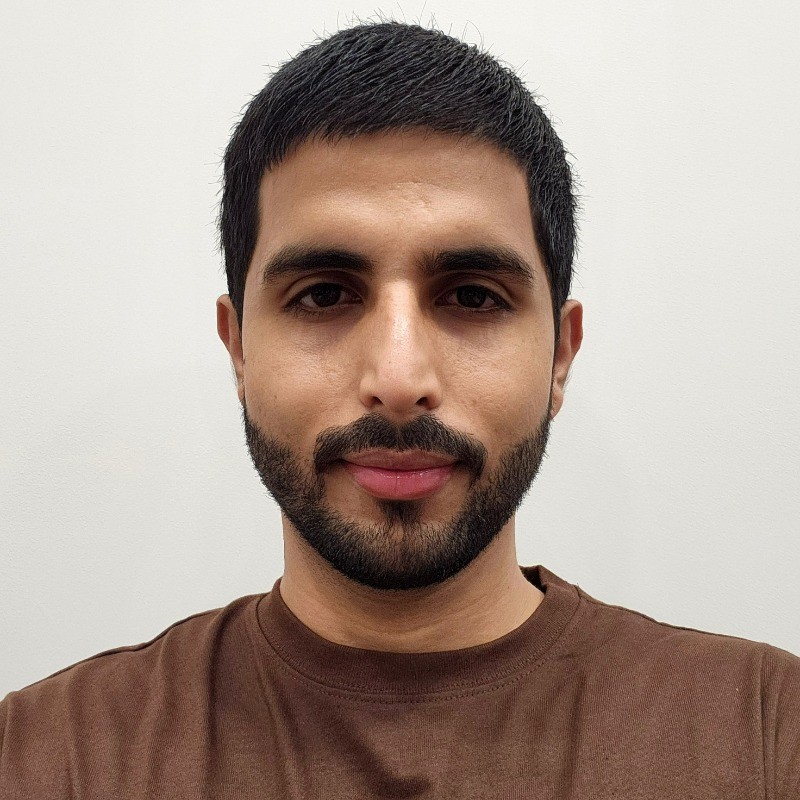 Portrait photograph of a man with dark hair and a full beard wearing a brown t-shirt against a neutral white background for professional headshot or identification purposes.